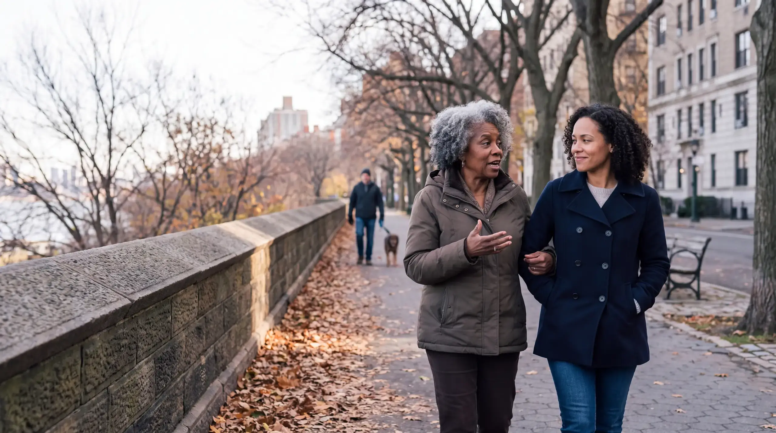 Older woman walking arm in arm with her adult daughter along a leafy New York park path in autumn, deep in conversation