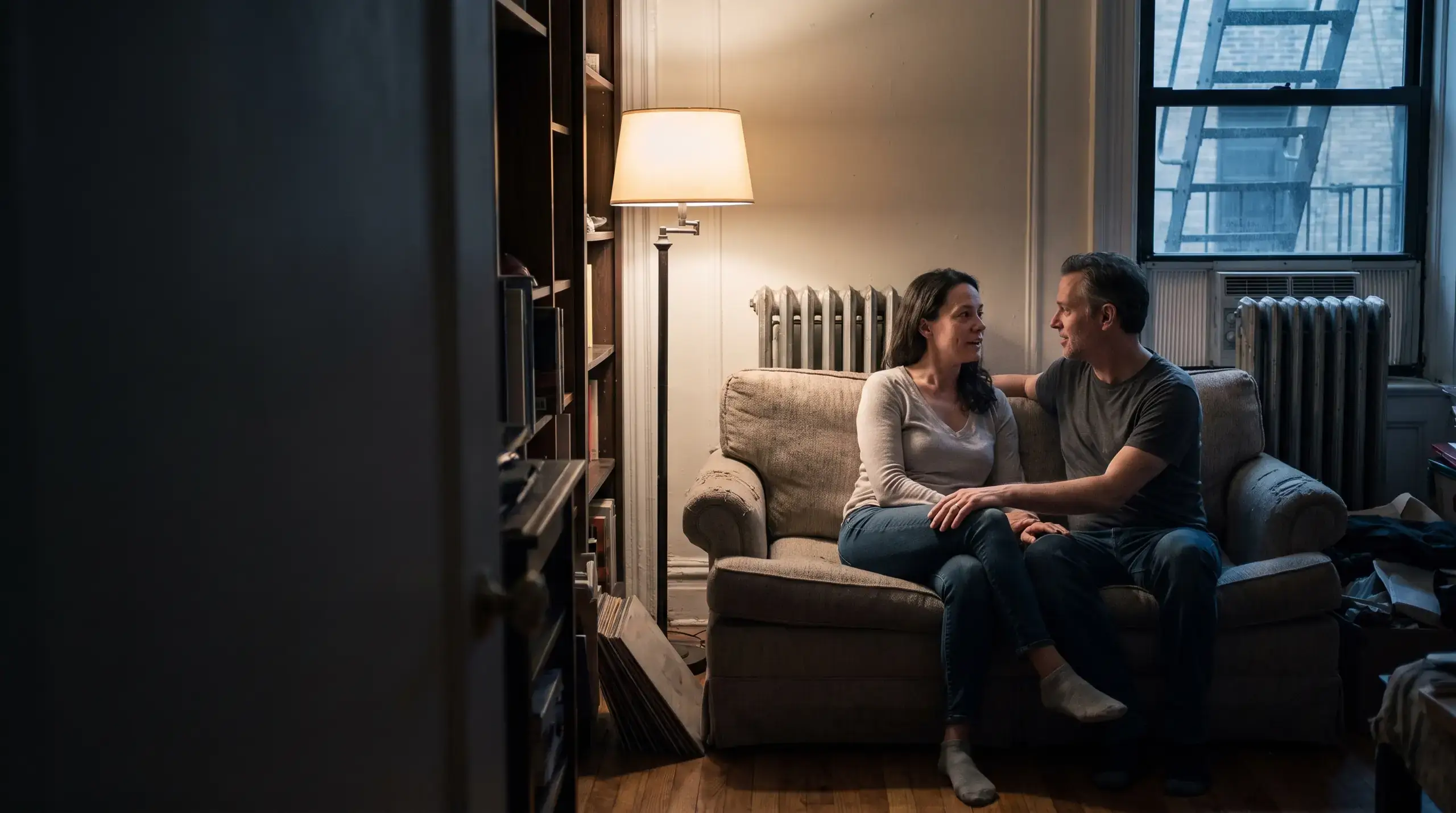 Couple sitting close on a sofa in lamplight, turned toward each other, talking things through